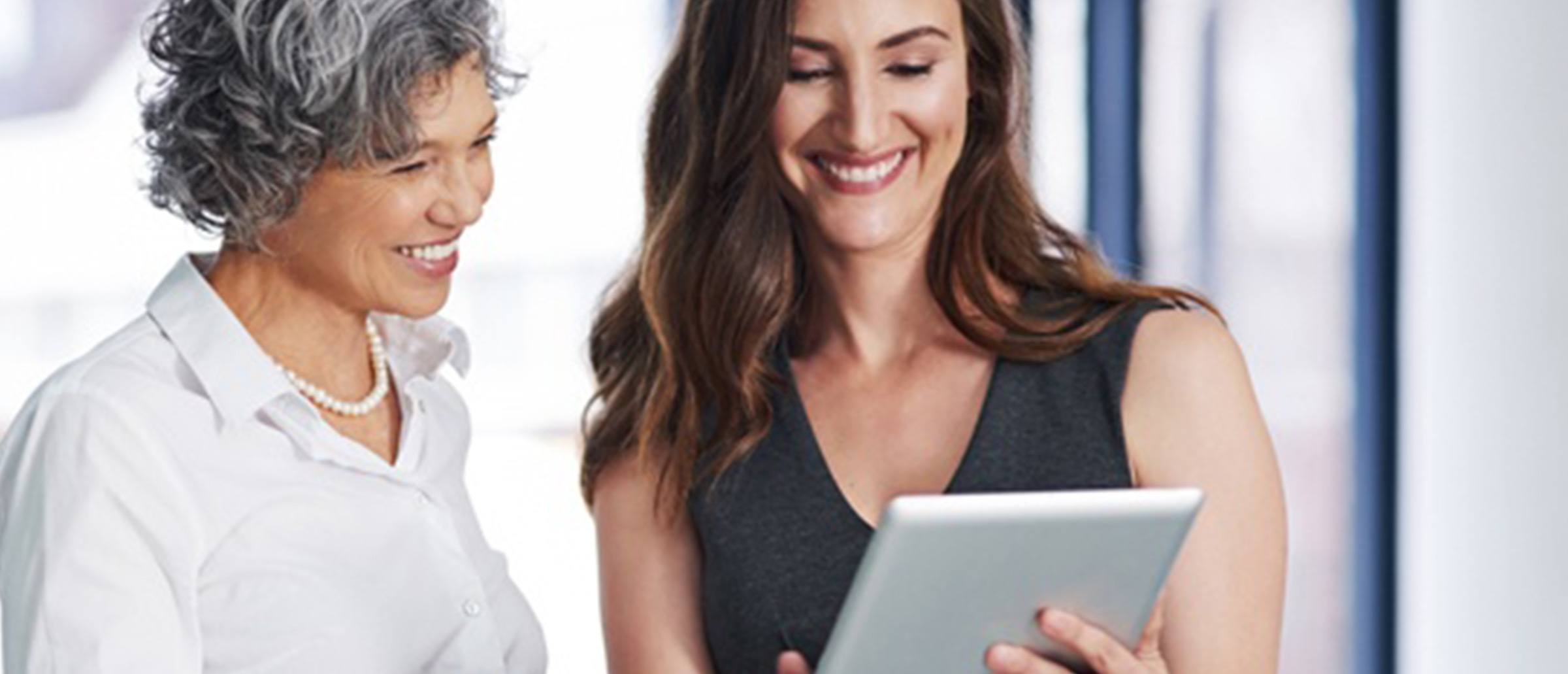 An older businesswoman and a younger businesswoman looking at a tablet device and smiling