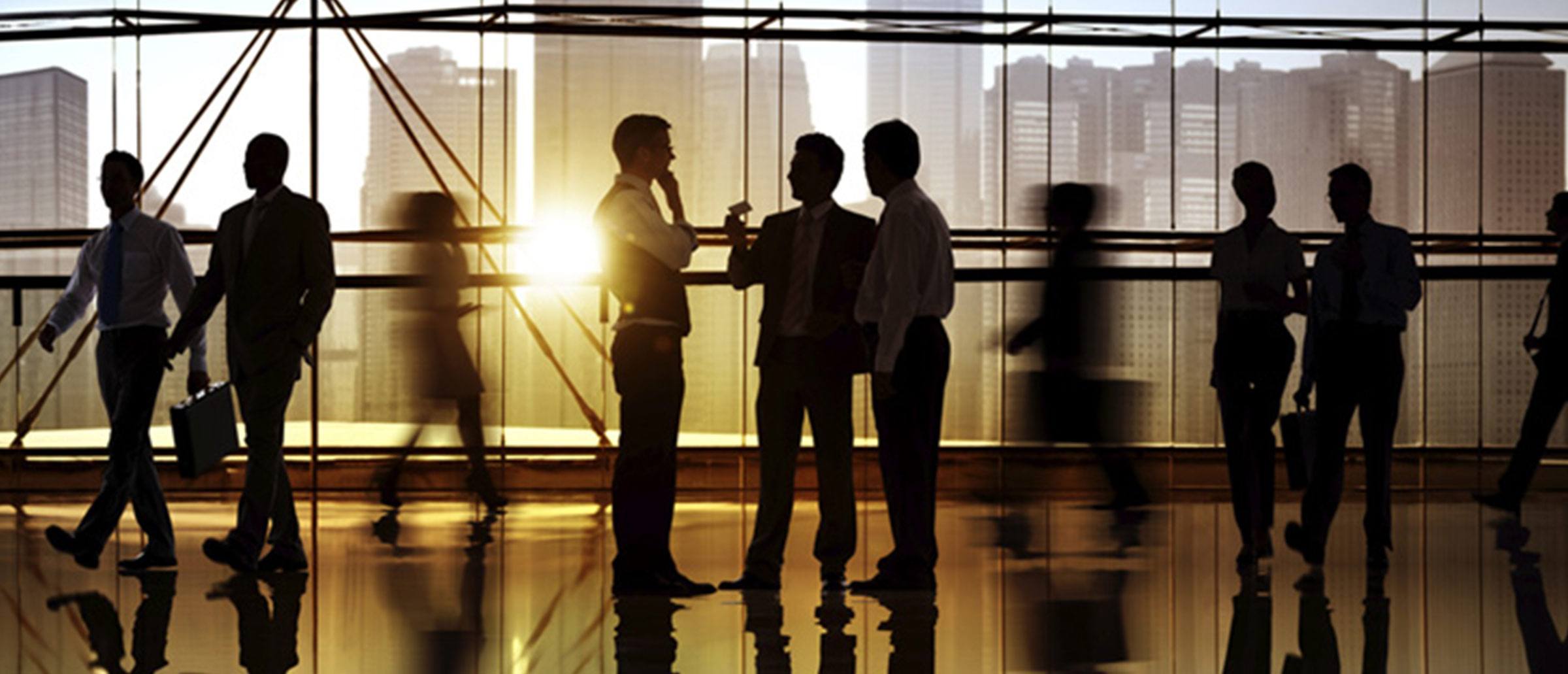 Dark hallway full of business people conversing and walking in front of a window overseeing a city skyline and sunset